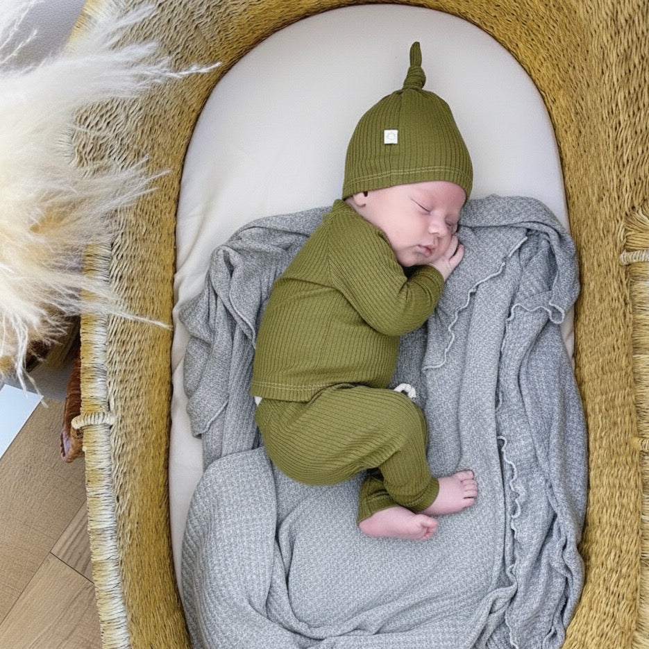 Baby in a green outfit and hat sleeping in a woven basket with a gray blanket.