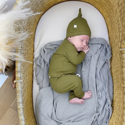 Baby in a green outfit and hat sleeping in a woven basket with a gray blanket.
