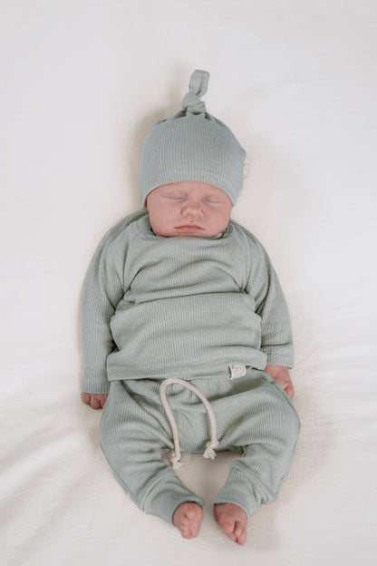 A sleeping newborn baby wearing a matching baby blue ribbed knit outfit, including a top knot hat and pants with a drawstring