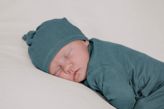 A sleeping newborn baby wearing a matching teal knit bonnet and waffle-knit bodysuit.
