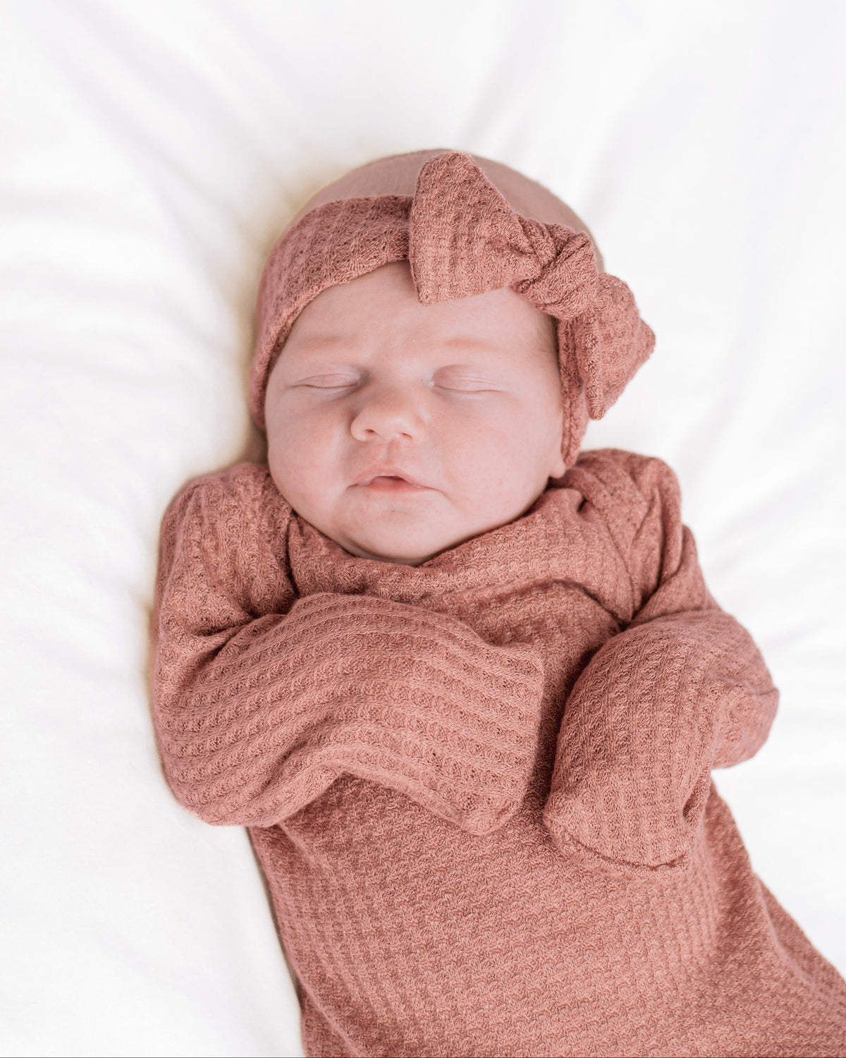 A sleeping newborn baby wrapped in a dusty rose waffle knit gown and matching bow headband