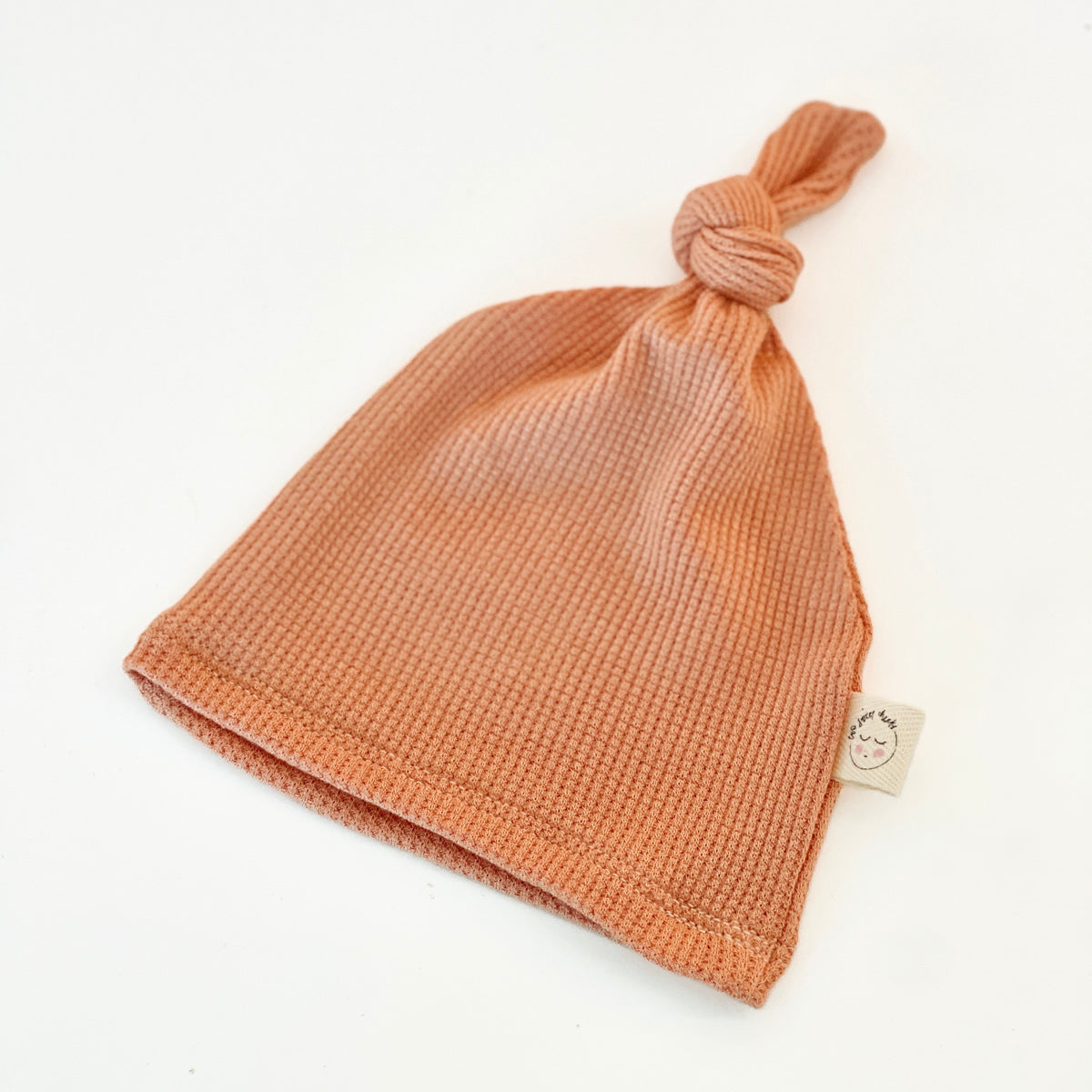 A knit baby beanie hat in a muted peach-orange color, featuring a knotted top