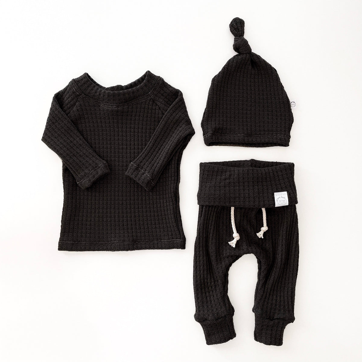 A three-piece baby knit clothing set in black waffle fabric, including a long-sleeve top, pants, and a knot  hat