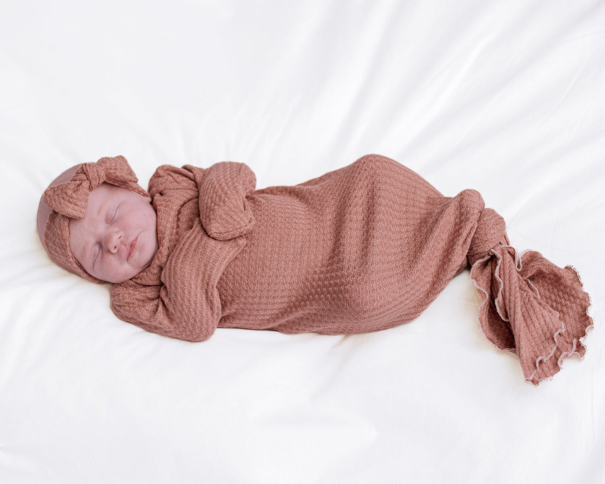 A sleeping newborn baby wrapped in a dusty rose waffle-knit gown with a matching knotted headband