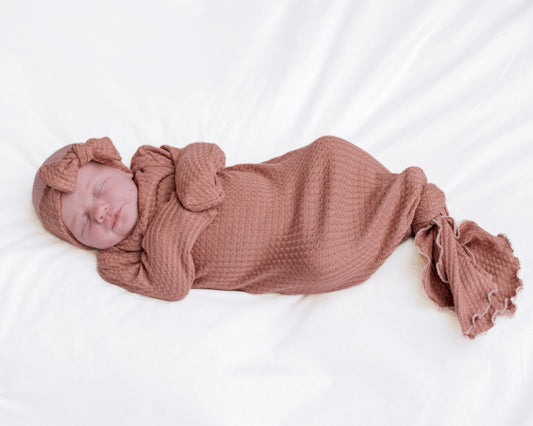 A sleeping newborn baby wrapped in a dusty rose waffle-knit gown with a matching knotted headband