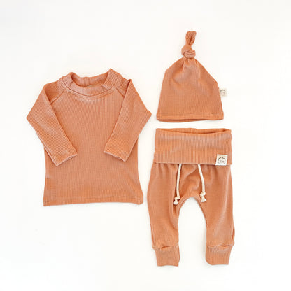 A three-piece baby clothing set in a warm terracotta or burnt orange waffle-knit fabric, including a long-sleeve top, pants with a drawstring, and a knotted beanie hat,