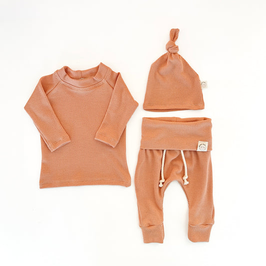 A three-piece baby clothing set in a warm terracotta or burnt orange waffle-knit fabric, including a long-sleeve top, pants with a drawstring, and a knotted beanie hat,
