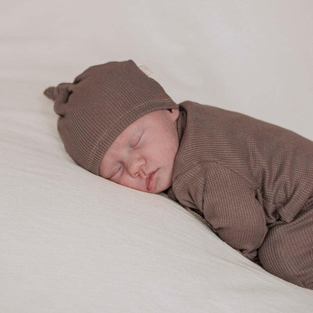 A sleeping newborn baby wearing a brown thermal knit hat and matching brown baby gown