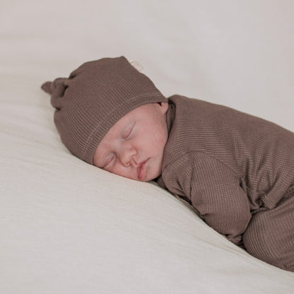 A sleeping newborn baby wearing a brown thermal knit hat and matching brown baby gown