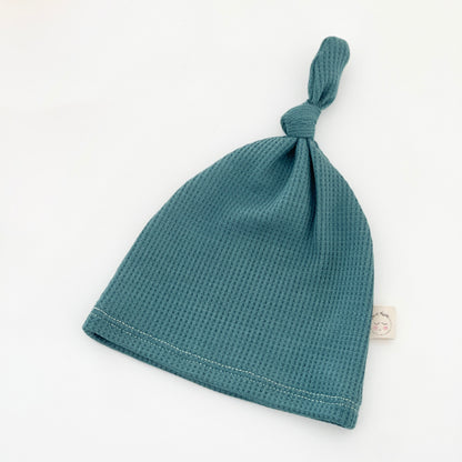 A teal blue-green waffle-knit baby beanie hat with a knotted top